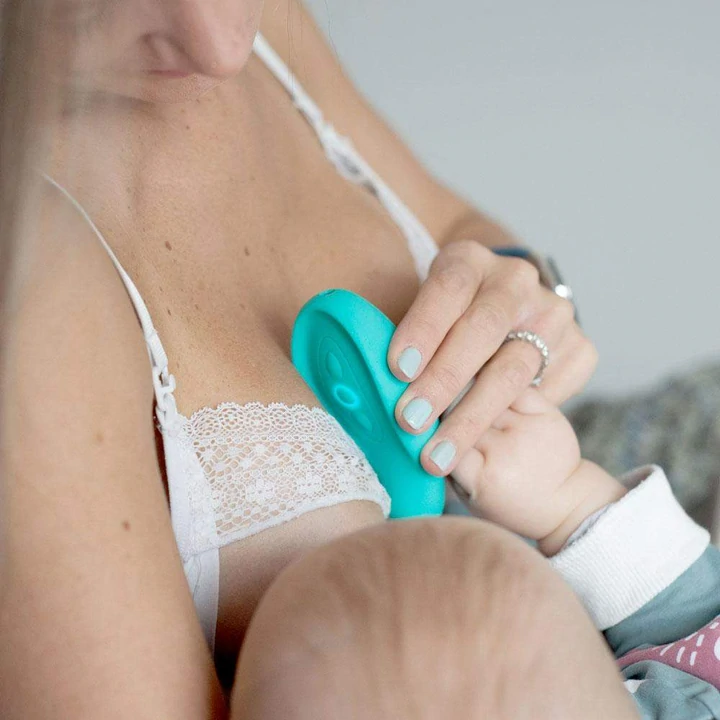 A woman breastfeeds her baby while using a blue lactation massager